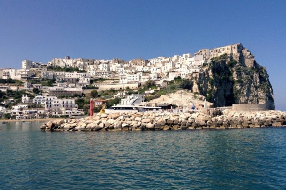 Vista panoramica della spiaggia di Peschici in estate, famiglie che giocano sulla sabbia, bambini con secchiello e paletta, mare cristallino sullo sfondo. Atmosfera rilassata e solare.